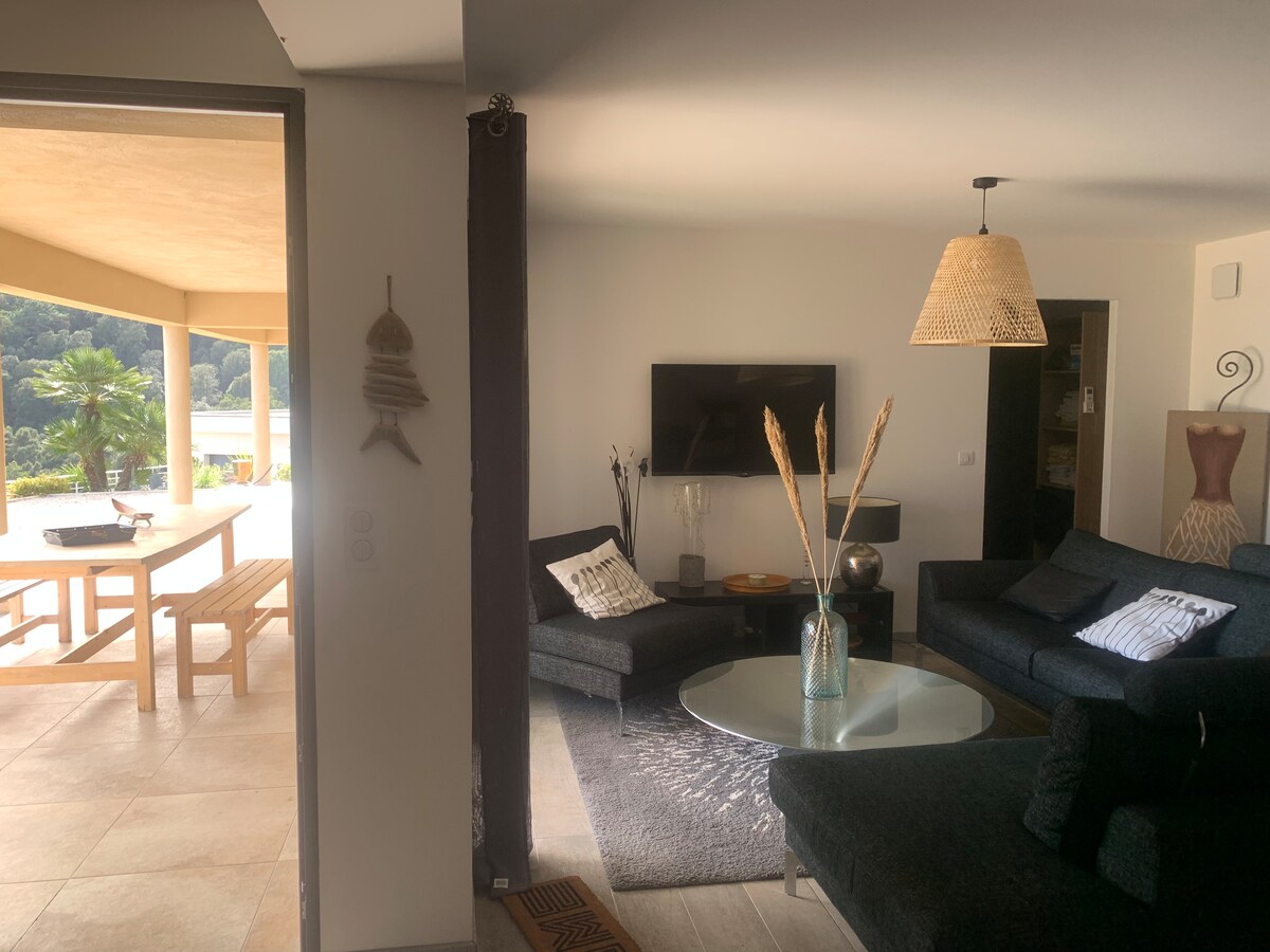 A spacious living area is depicted, featuring comfortable dark sofas arranged around a glass coffee table. A large television is mounted on the wall, while a stylish pendant light hangs above. Light from the exterior illuminates the room, enhancing the serene atmosphere.