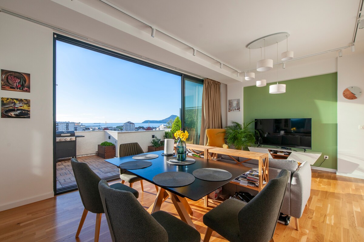 A spacious living area features a modern dining table surrounded by gray chairs. A large window provides a view of the sea and mountains. A television is mounted on a green accent wall, while natural light enhances the warm wood flooring.