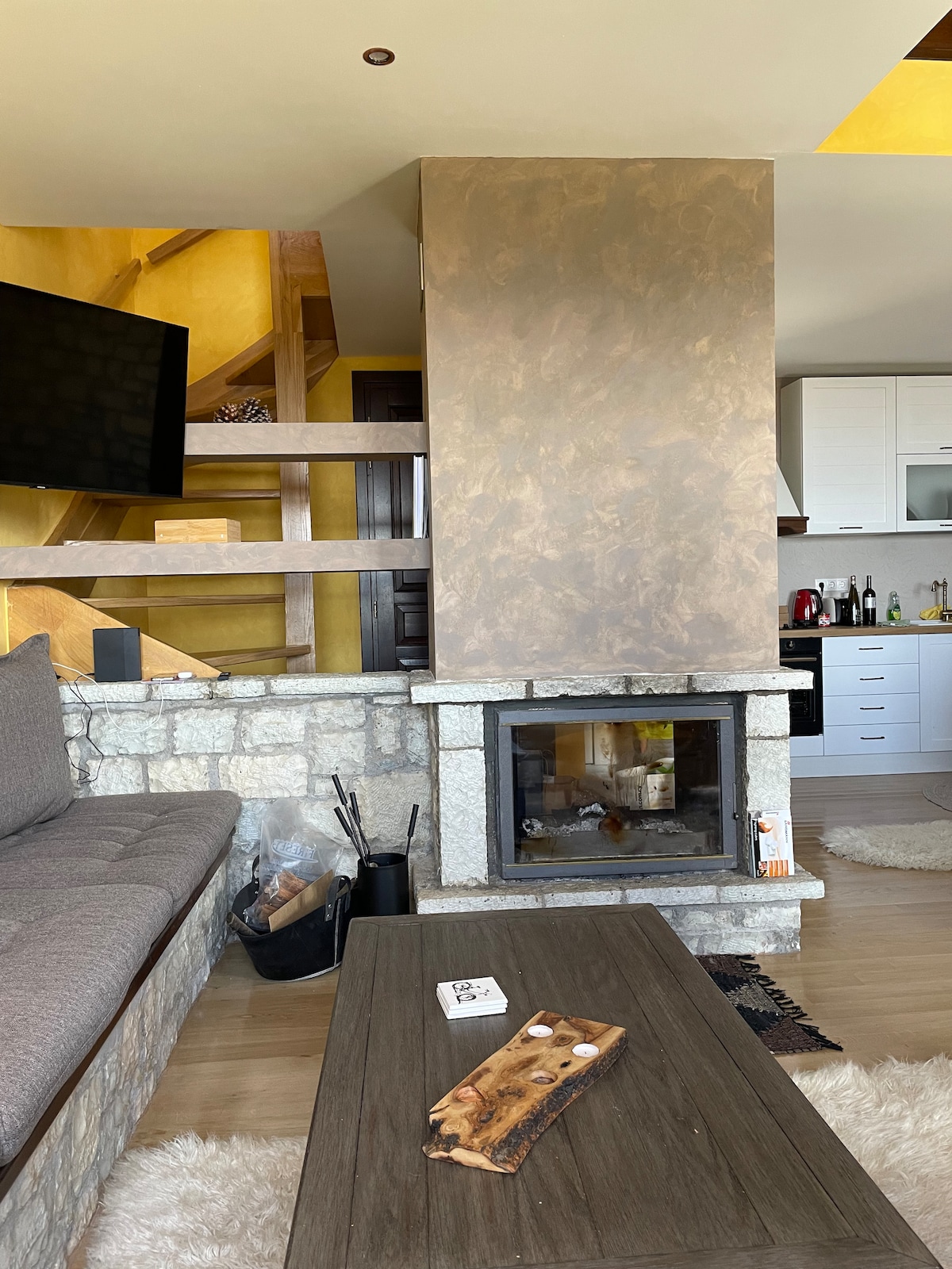 A cozy living area features a stone fireplace integrated into a wooden wall. A contemporary coffee table, adorned with decorative items, sits in front of a plush sofa. A large television is mounted on the wall, and a fully equipped kitchen is visible in the background.