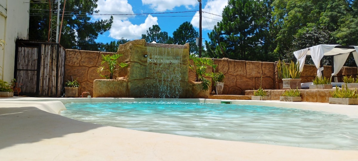 A serene pool area is featured, showcasing clear water with a gentle waterfall flowing into the pool. Surrounding the pool are lush green plants and relaxing lounge spaces, all set against a tranquil backdrop of trees and a partly cloudy sky.