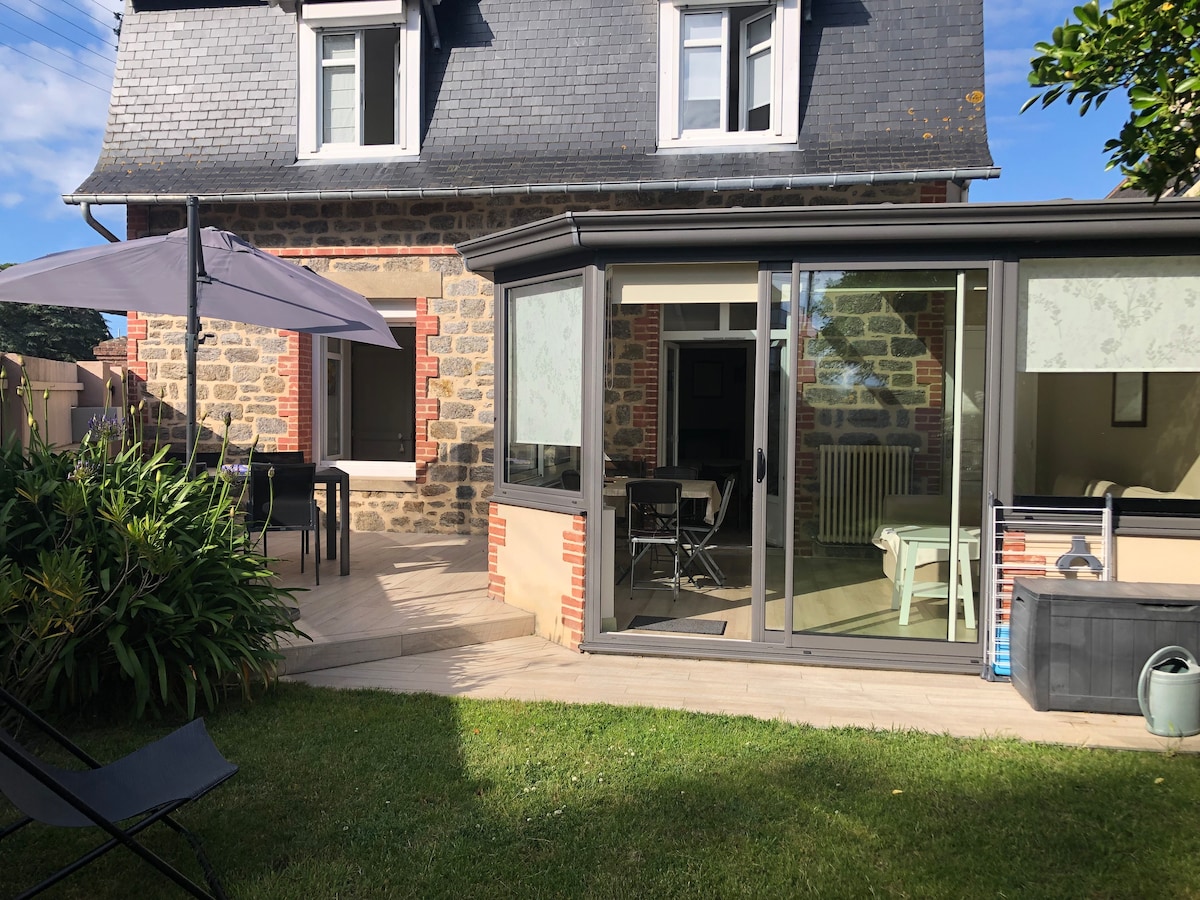 The exterior view showcases a charming stone house with a spacious patio area, featuring a glass-enclosed veranda and outdoor furniture. A green lawn surrounds the space, complemented by an awning and a decorative plant border. Clear skies are visible above, enhancing the inviting atmosphere.