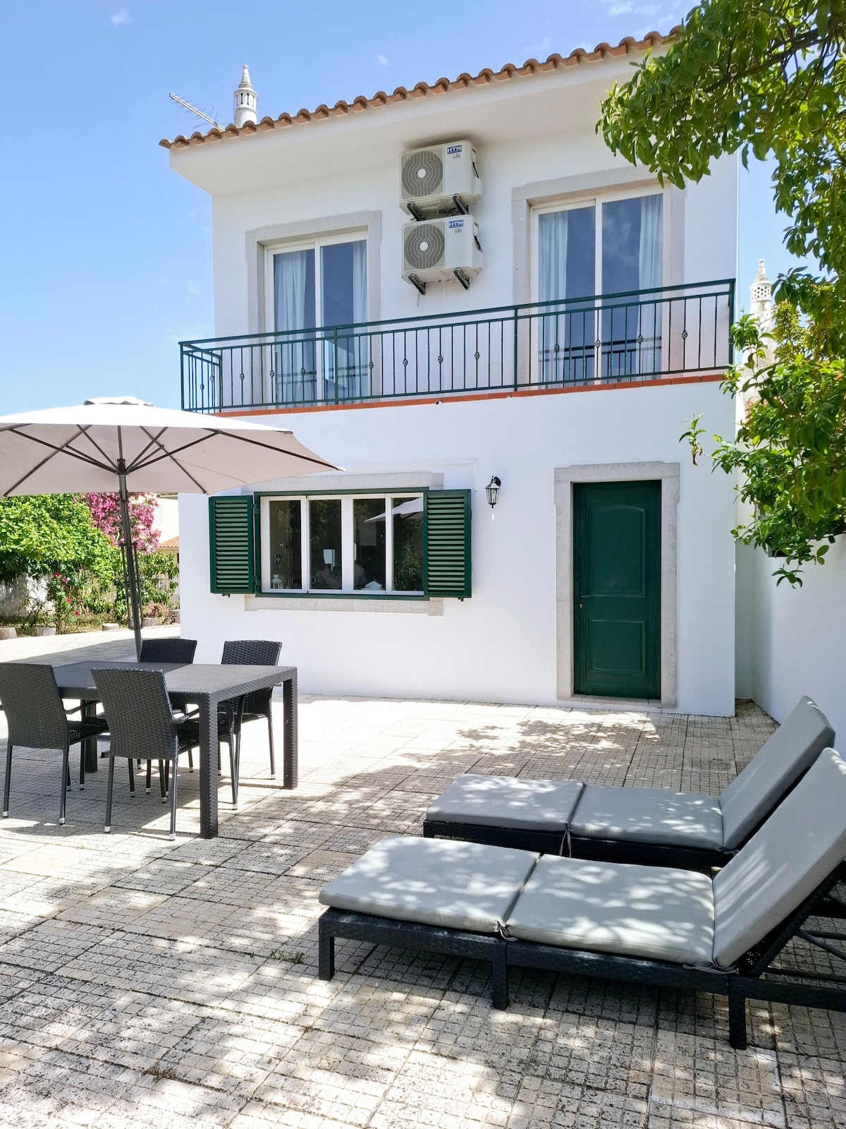 An outdoor area features a dining table for six and two lounge chairs, positioned on a tiled patio. A large umbrella provides shade over the dining space. Green shutters accent the white exterior of the two-story building, which includes air conditioning units visible on the upper level.