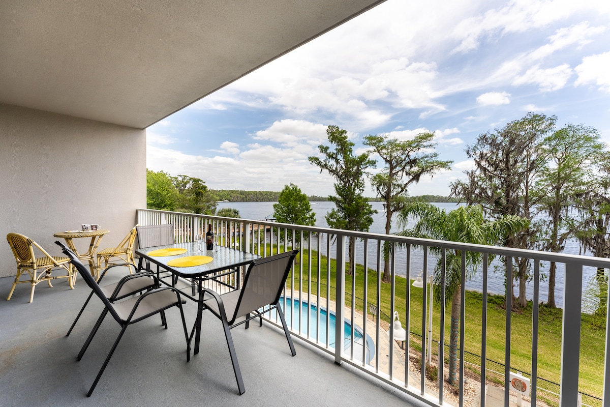 A large private balcony overlooks Lake Bryan, featuring a dining table with yellow placemats and four chairs. Two loungers provide a space for relaxation. The tranquil waters and lush greenery create a serene setting, while a pool is visible just below.