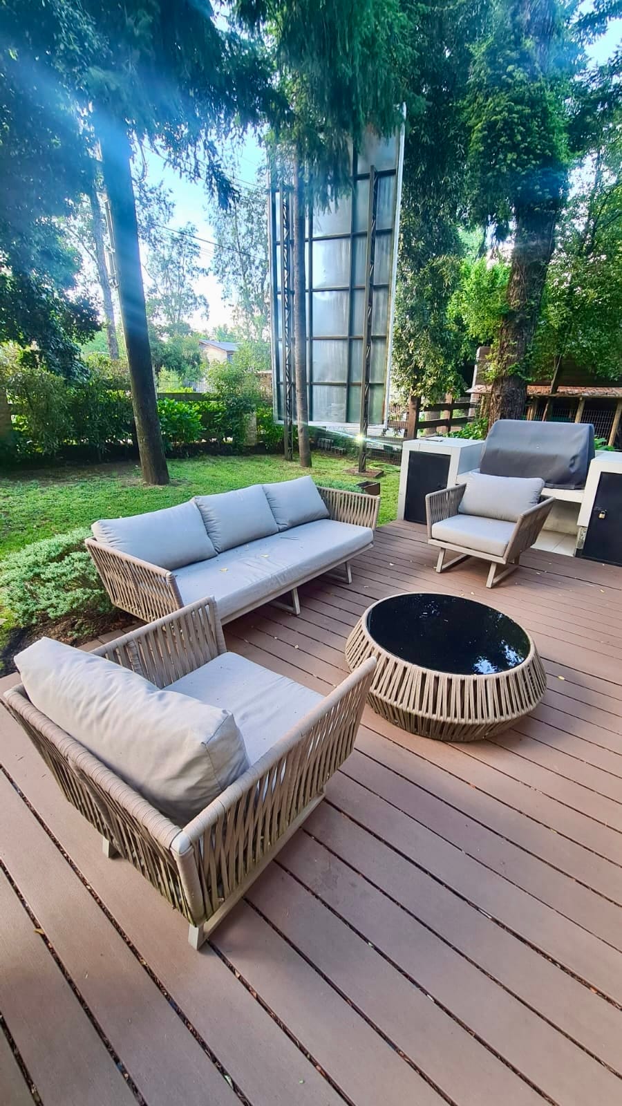 An outdoor seating area features a round coffee table surrounded by a spacious sofa and two armchairs with soft cushions. The deck has a wood-like finish and is enclosed by lush greenery, creating a serene atmosphere for relaxation.