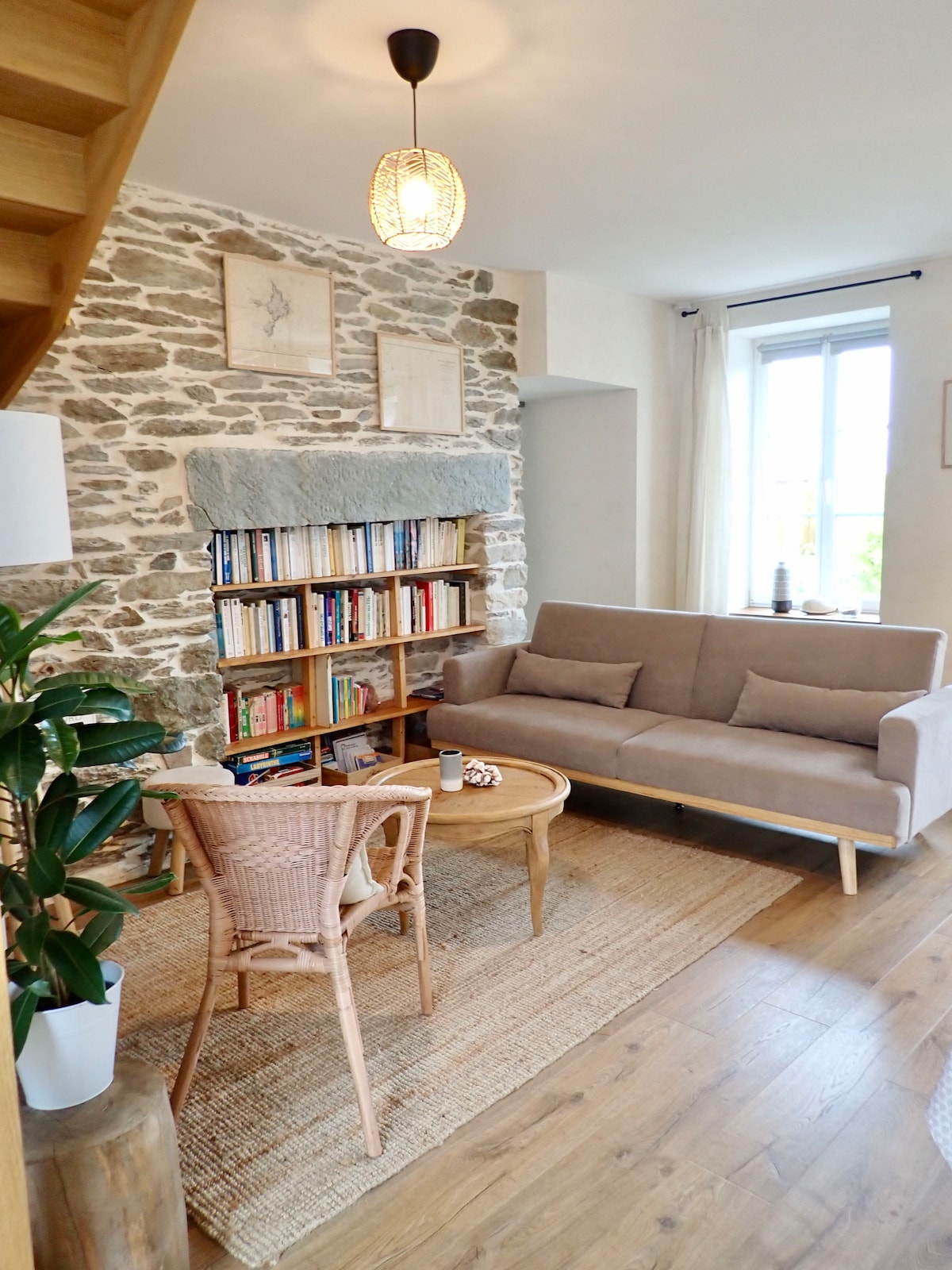 A welcoming living area is highlighted by a stone wall and a soft sofa facing a wooden coffee table. A bookshelf filled with various books offers a cozy touch. Natural light enters through the window, complemented by a simple decorative plant nearby.