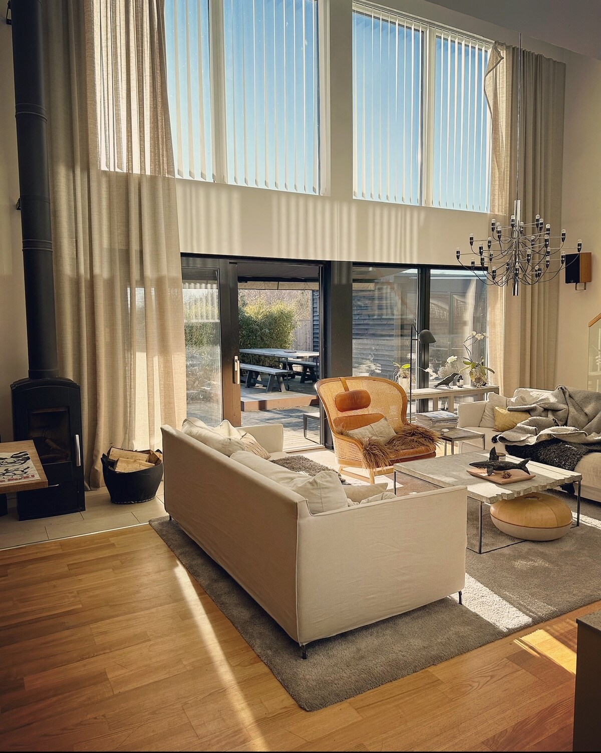 A spacious living area features large glass sliding doors that open to an outdoor space. Tall windows allow natural light to fill the room, highlighting a mix of modern furniture, including a comfortable orange chair and a light-colored sofa. The wooden floor and soft area rug add warmth.