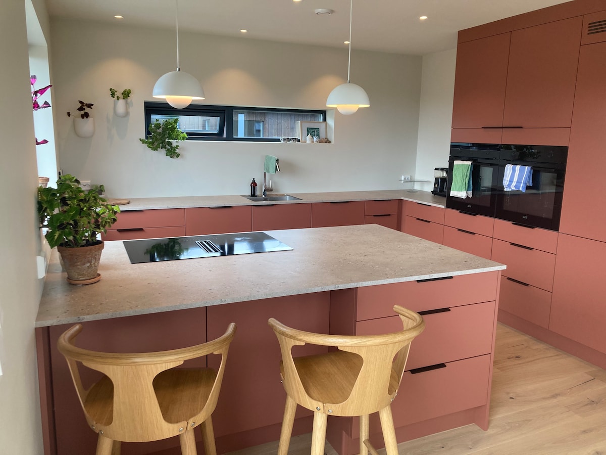 A modern kitchen features soft-toned cabinetry and a central island with an integrated cooktop. Two wooden stools are positioned at the island. Large windows allow natural light to fill the space, complemented by minimal wall decor and various plants enhancing the atmosphere.