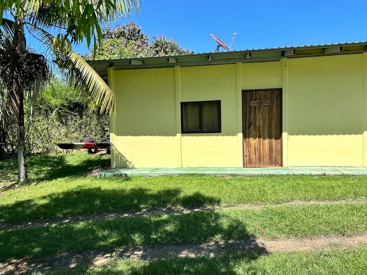 A single-story yellow building with a wooden door and a small window is visible. The structure is surrounded by green grass, with a palm tree nearby. A kayak is partially shown in the background, suggesting outdoor activities are available.