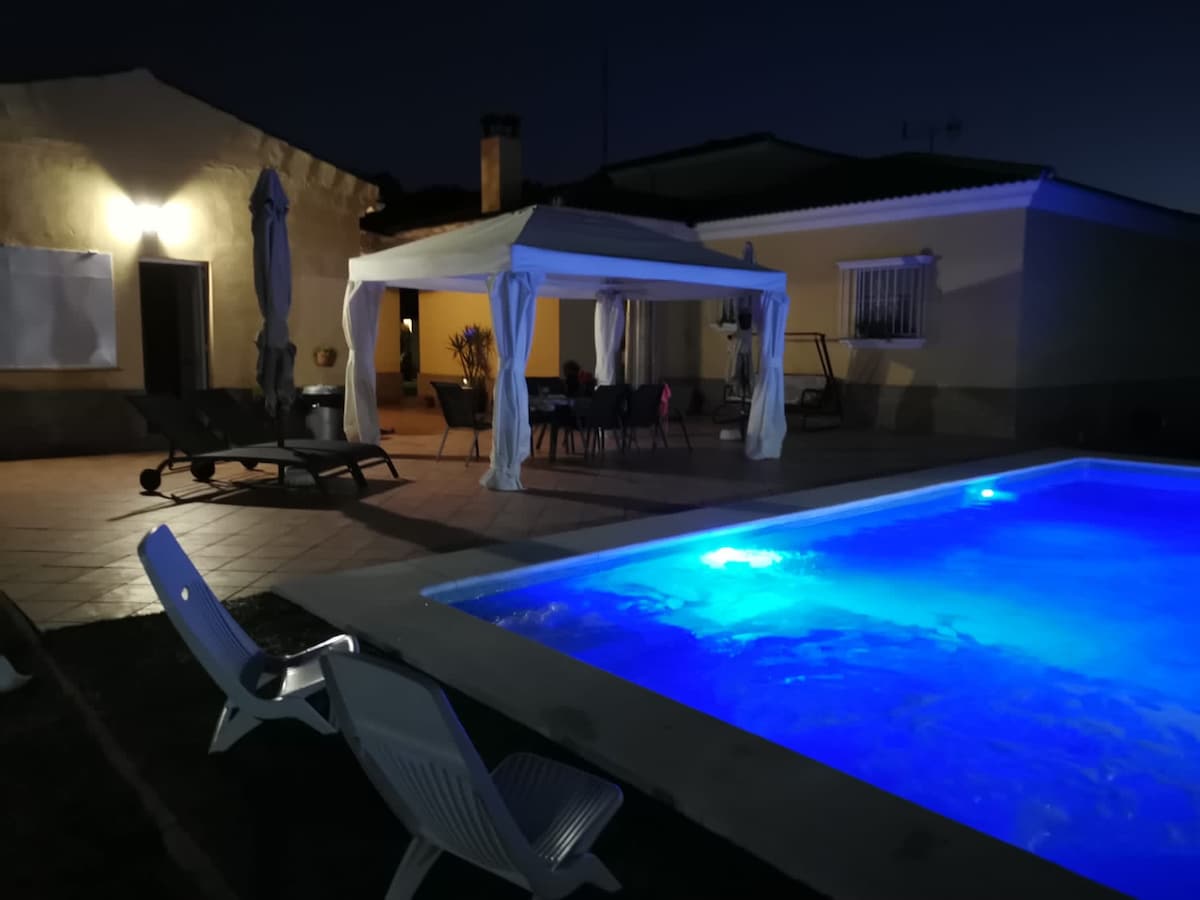 At night, the illuminated pool glows a tranquil blue, while a shaded terrace area features seating and a dining table under a canopy. Lounge chairs are positioned nearby, surrounded by a peaceful outdoor setting.