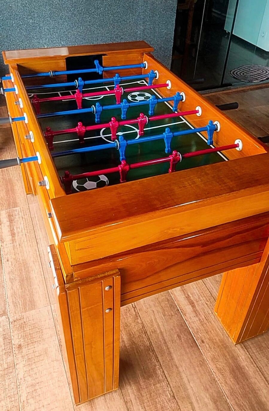 A wooden foosball table is displayed, featuring bright blue and pink players arranged on a green playing field with white markings. The table's smooth surface reflects the surrounding light, while its sturdy design is evident in the thick, polished legs.