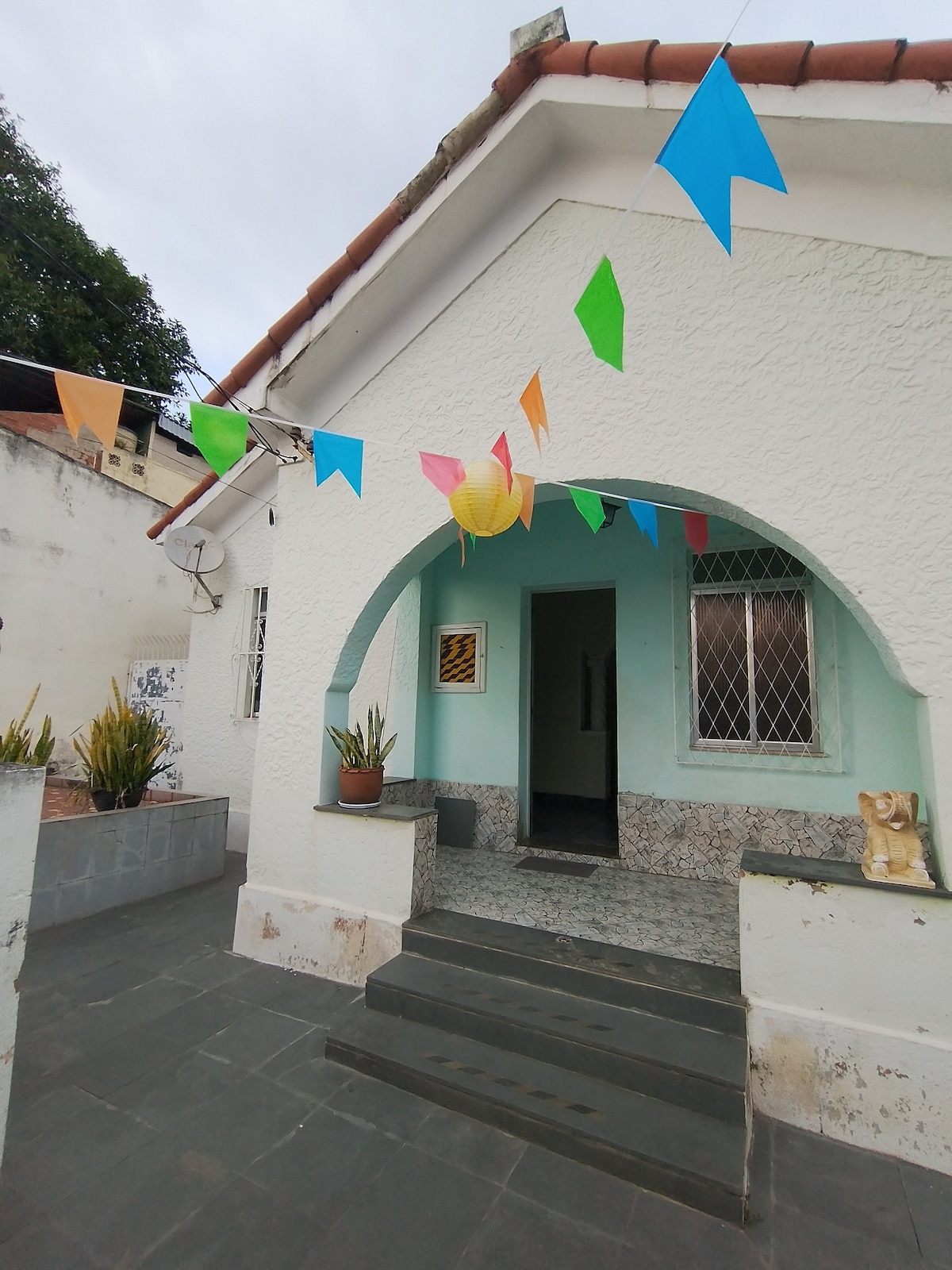 The entrance features an arched doorway leading into a light blue facade, creating a cheerful ambiance. Colorful banners hang above the entryway, complemented by potted plants on a stone step. The floor is lined with dark tiles, enhancing the welcoming appearance of the home.