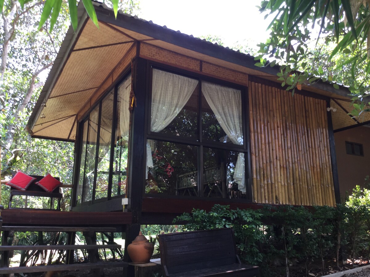 A charming cabin features large windows framed by sheer curtains, allowing natural light to fill the space. The exterior showcases a mix of bamboo and wood materials, complemented by a small deck with seating and surrounding greenery.