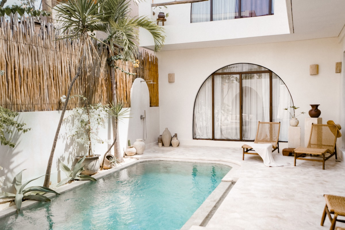 A serene outdoor area is highlighted by a refreshing pool surrounded by natural elements. Wooden lounge chairs are positioned nearby, and a tall palm tree adds an inviting touch. The space is framed by a textured wooden fence and an arched window, creating a tranquil atmosphere.
