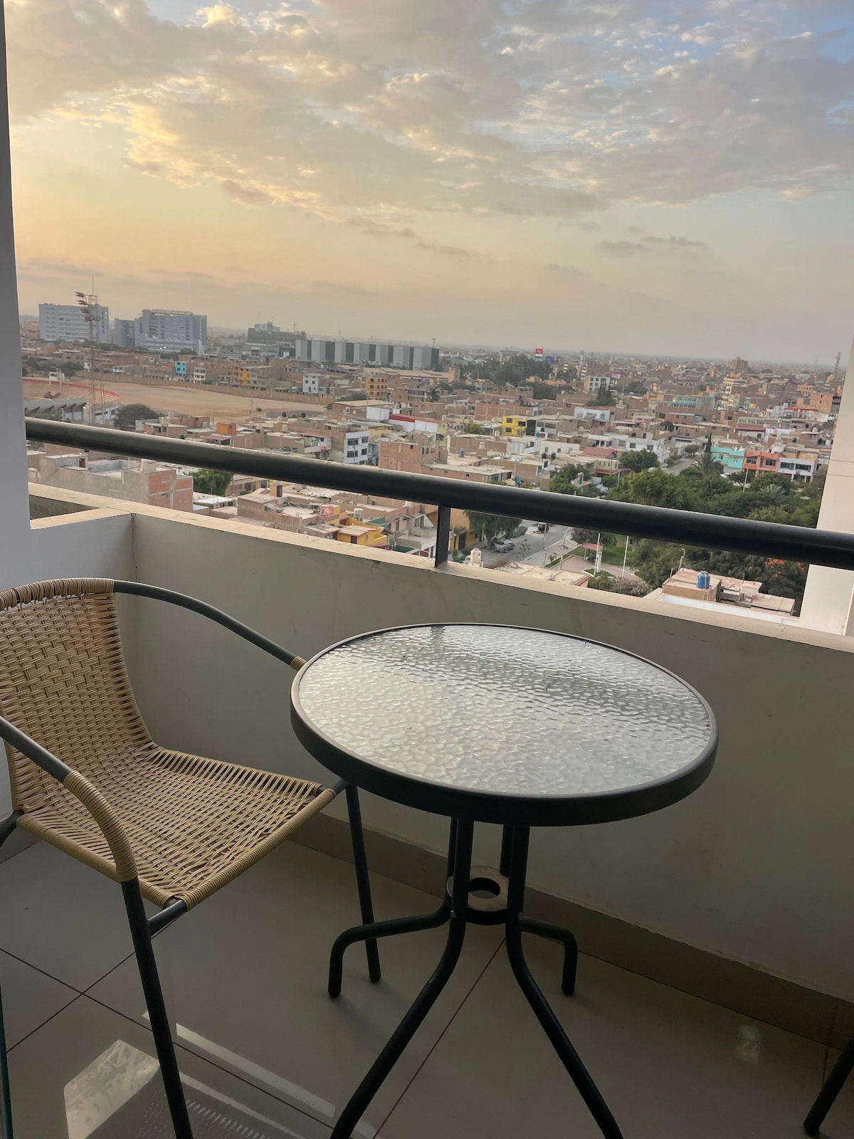 A balcony features a round table with a textured surface and a single wicker chair, offering a vantage point of the surrounding cityscape. The horizon displays a blend of buildings under a lightly clouded sky, with warm hues from the setting sun illuminating the scene.