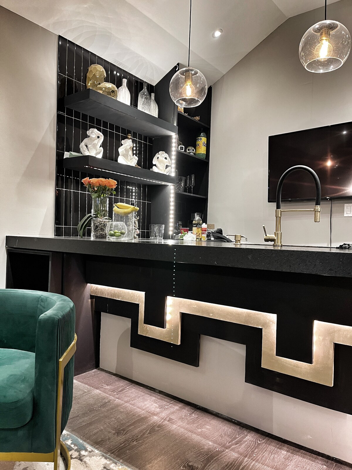 A modern bar area features a sleek dark countertop, outlined by decorative lighting in a zigzag pattern. Open shelves display a variety of glassware and decorative sculptures. A stylish green chair provides seating, with pendant lights illuminating the space above.