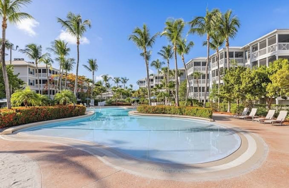 An inviting outdoor lagoon pool is surrounded by lush tropical landscaping and tall palm trees. A spacious deck with sun loungers provides ample space for relaxation. The pool area is well-maintained, showcasing clear blue water under bright skies.