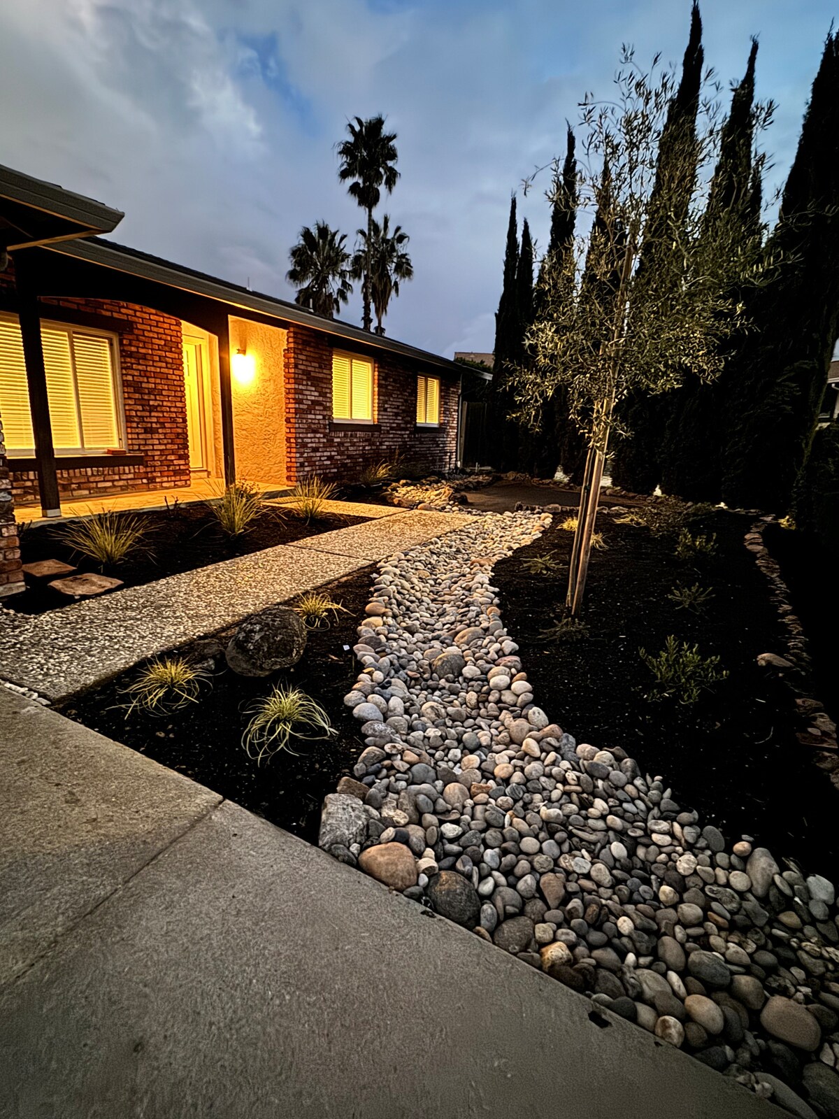 The exterior of the modern farmhouse features a welcoming pathway lined with smooth stones, guiding the way to the entrance. Lush low-maintenance landscaping with palm and cypress trees creates a serene ambiance, enhanced by soft illumination from exterior lighting.
