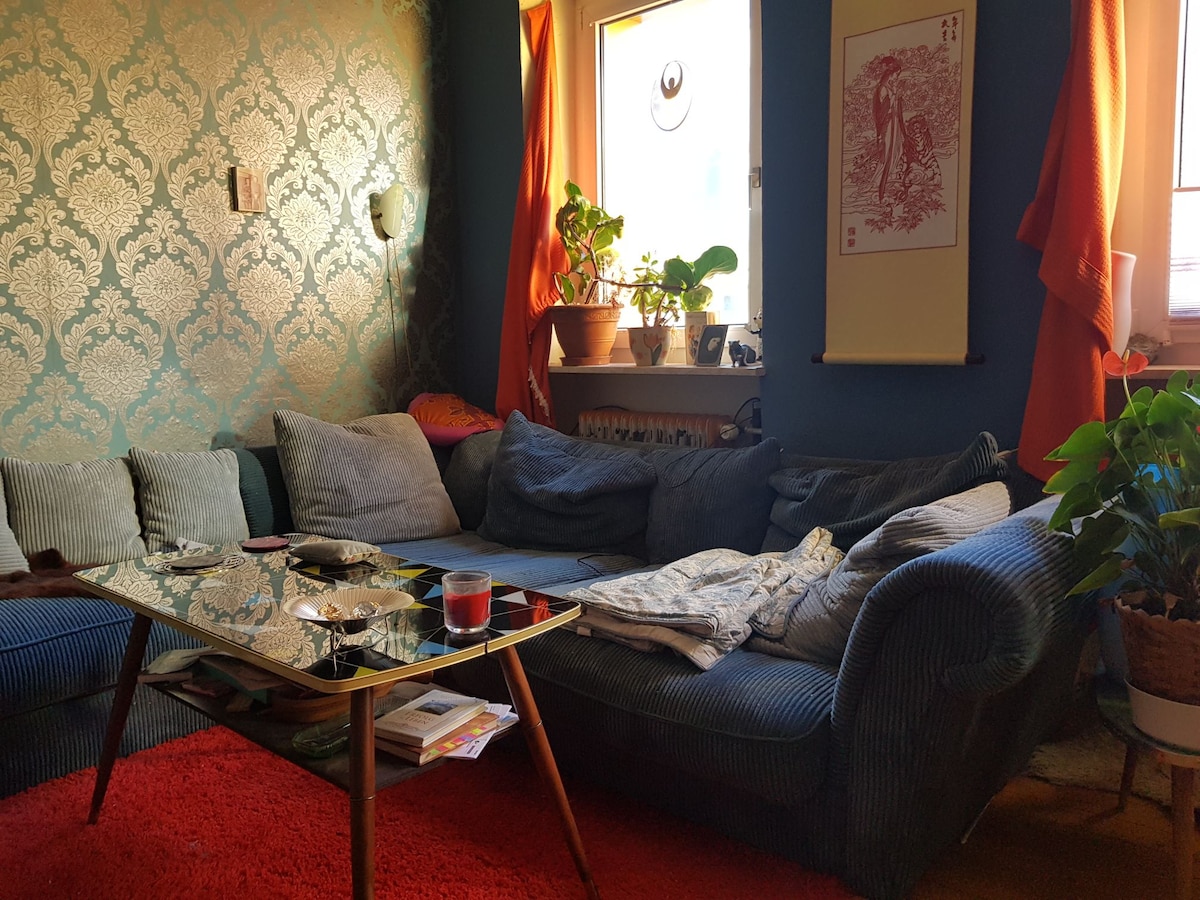 A seating area features a large, plush sofa adorned with decorative pillows. A low coffee table sits in front of the sofa, complemented by plants and artistic wall prints. Natural light enters through a window, enhancing the vibrant colors of the room.