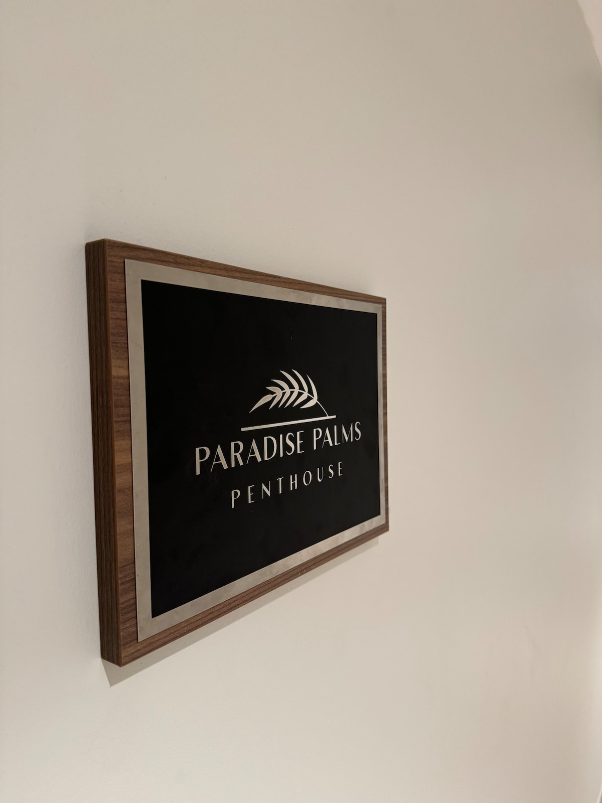 A framed sign is displayed on a light-colored wall, indicating the name 'Paradise Palms Penthouse.' The sign features a simple black background with white lettering and a minimalistic design that includes a palm leaf motif.