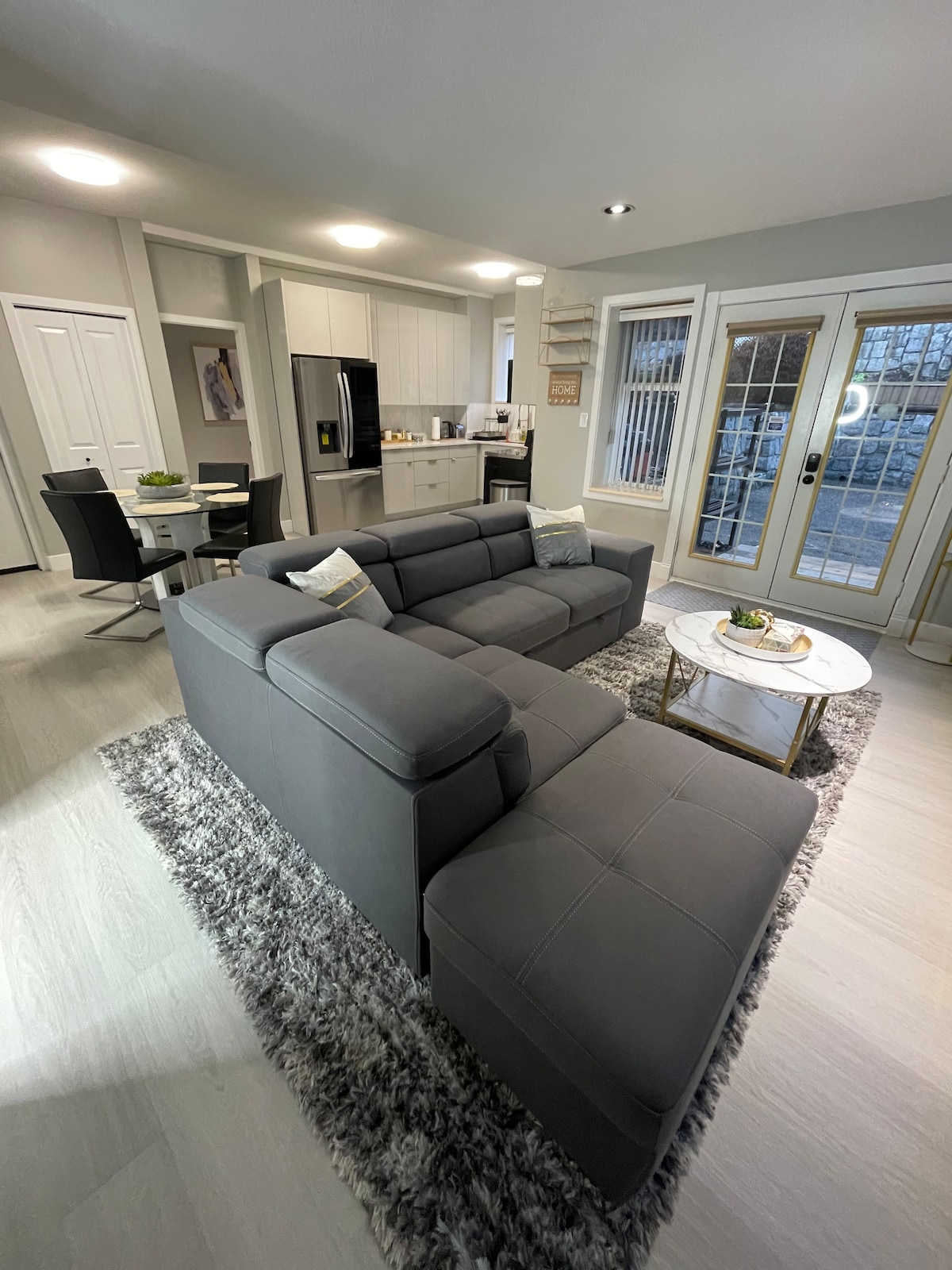 A modern living area is defined by a spacious gray sectional sofa facing a sleek coffee table. Natural light filters through large glass doors that lead to the outdoor space. An open kitchen with contemporary appliances is visible in the background, accompanied by a dining area with elegant chairs.