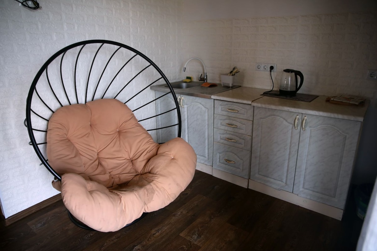 A cozy corner is presented with a large, soft hanging chair featuring plush cushions. Nearby, a small kitchenette includes a kettle, sink, and counter space, positioned against a textured white wall. The floor is finished with dark wood, adding warmth to the area.