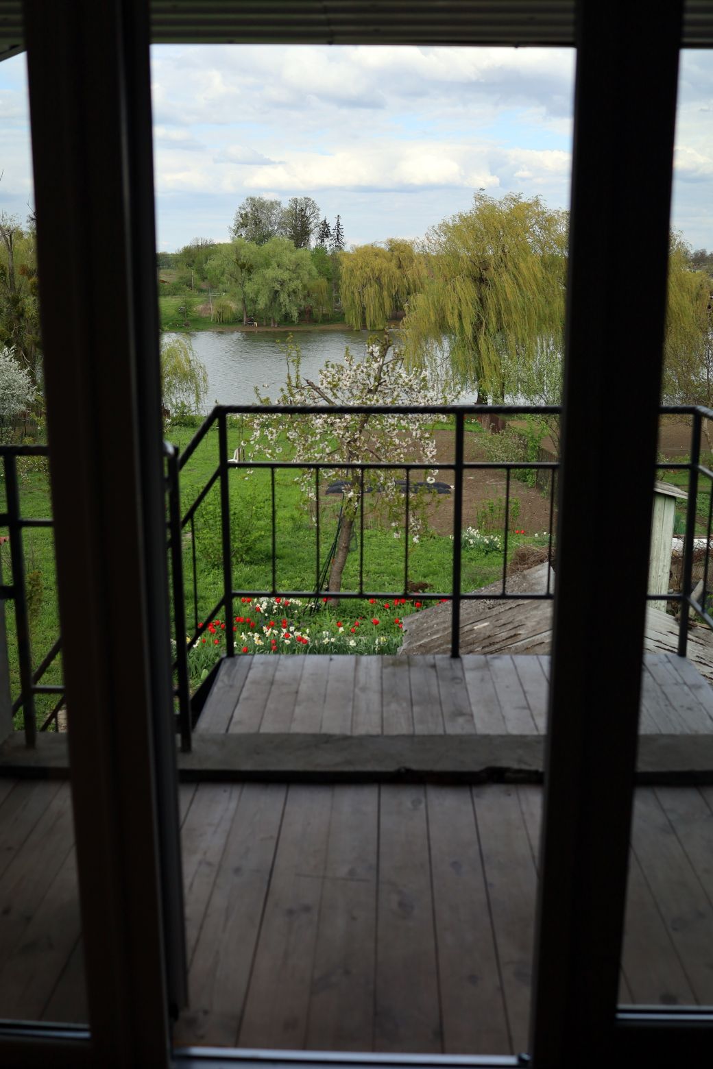 A wooden balcony extends from the living area, offering a view of a serene lake surrounded by lush greenery and flowering trees. The scene is framed by glass doors, highlighting the natural beauty of the outdoor space.