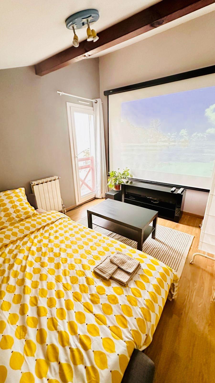 A cozy sleeping area is featured, with a spacious bed dressed in a yellow polka-dotted duvet. A small coffee table sits in front, accompanied by a plant. Natural light enters through the window, illuminating the room's neutral walls and hardwood floor.