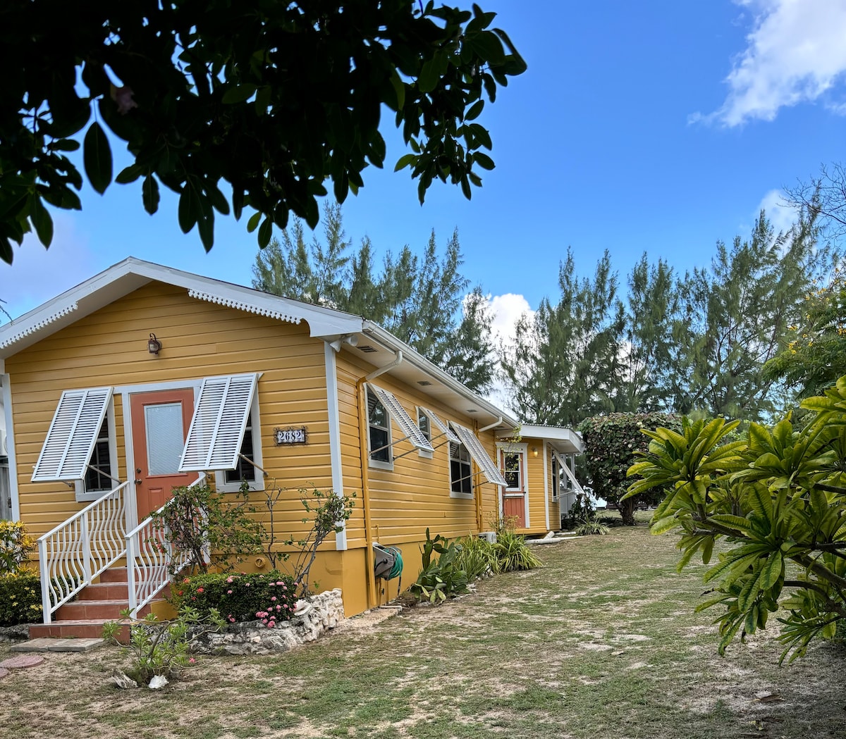 A charming yellow bungalow is surrounded by lush greenery and blue skies. White shutters adorn the windows, while vibrant plants and trees frame the property. A landscaped yard provides an inviting outdoor space, reflecting a tranquil Caribbean atmosphere.