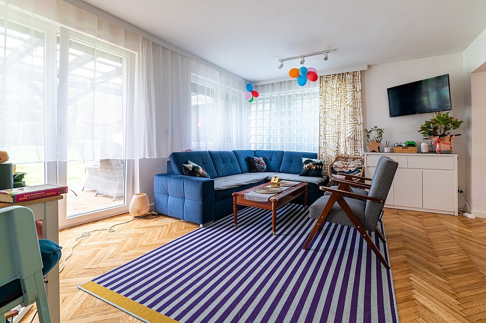 A spacious living area features a blue sectional sofa and two wooden armchairs, arranged around a low coffee table. A striped area rug enhances the space, while large glass doors lead to the outside terrace. Soft natural light fills the room, complemented by colorful decor.