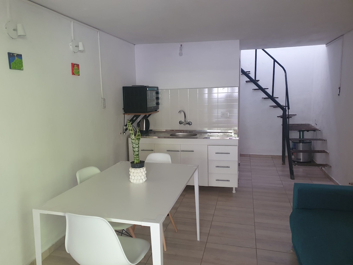 An open kitchen area is paired with a dining table set for four. Modern white cabinetry complements the sleek stainless steel sink. A spiral staircase leads to the upper level, while natural light enhances the spacious layout.