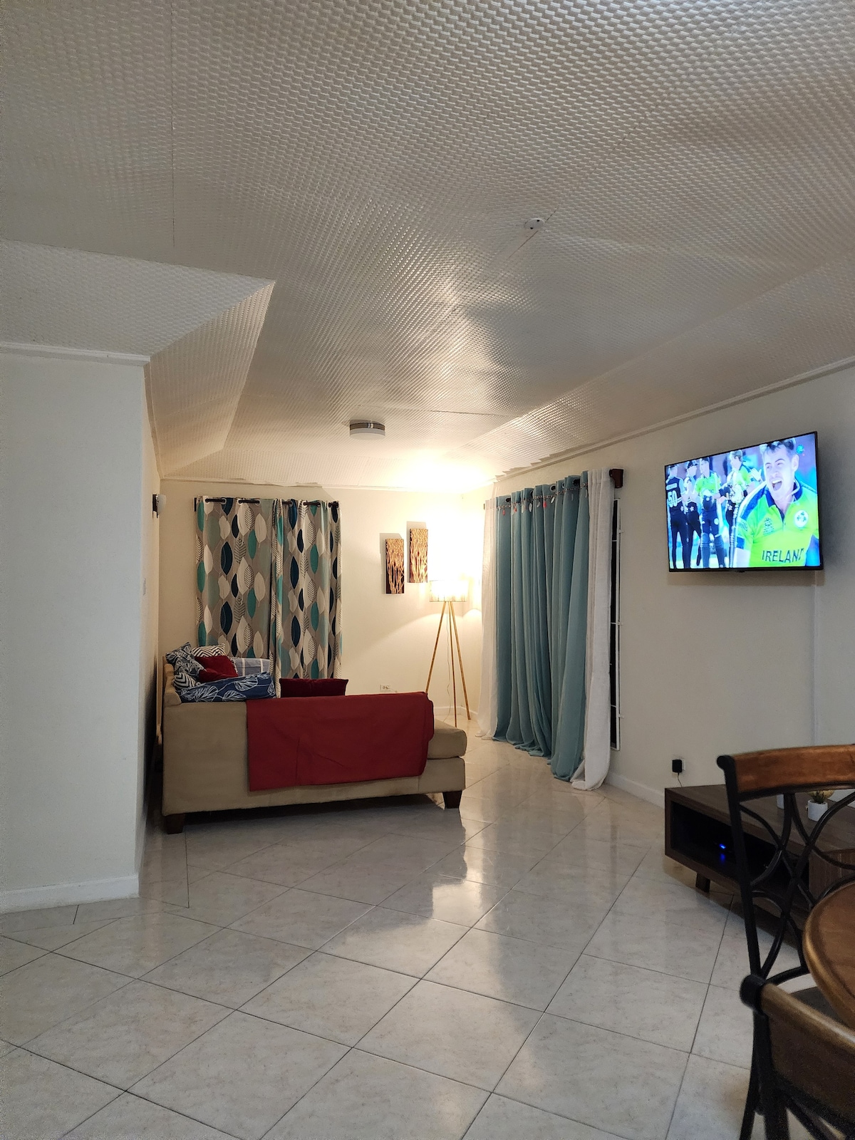 A living room area is observed featuring a comfortable sofa adorned with a red throw. Soft, patterned curtains frame a window, while a floor lamp provides warm light. A television mounted on the wall offers entertainment options, and the tiled floor reflects light, enhancing the spacious feel.