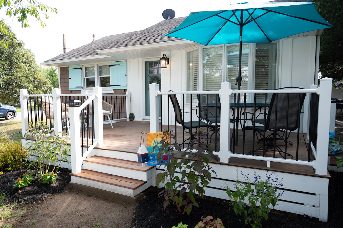 A front porch is depicted with a dining table and chairs underneath a large turquoise umbrella. Steps lead down to a landscaped area with various plants. The exterior features white railings and a door with window shutters, creating a welcoming entryway.