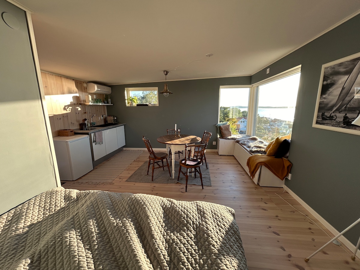A bright studio space features a well-equipped kitchen area and a cozy dining table surrounded by chairs. Large windows allow natural light to illuminate the room while offering views of the sea. A comfortable nook with cushions invites relaxation.