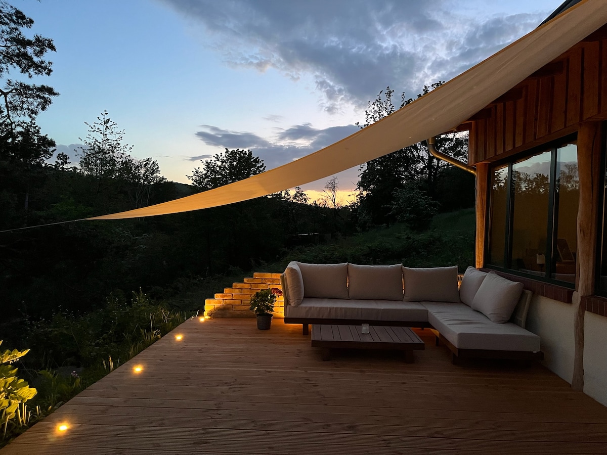 A spacious wooden terrace is seen under a soft evening sky, illuminated by subtle LED lights. A comfortable outdoor seating area is furnished with plush cushions and a low coffee table. A small potted plant adds a touch of greenery, enhancing the natural ambiance.