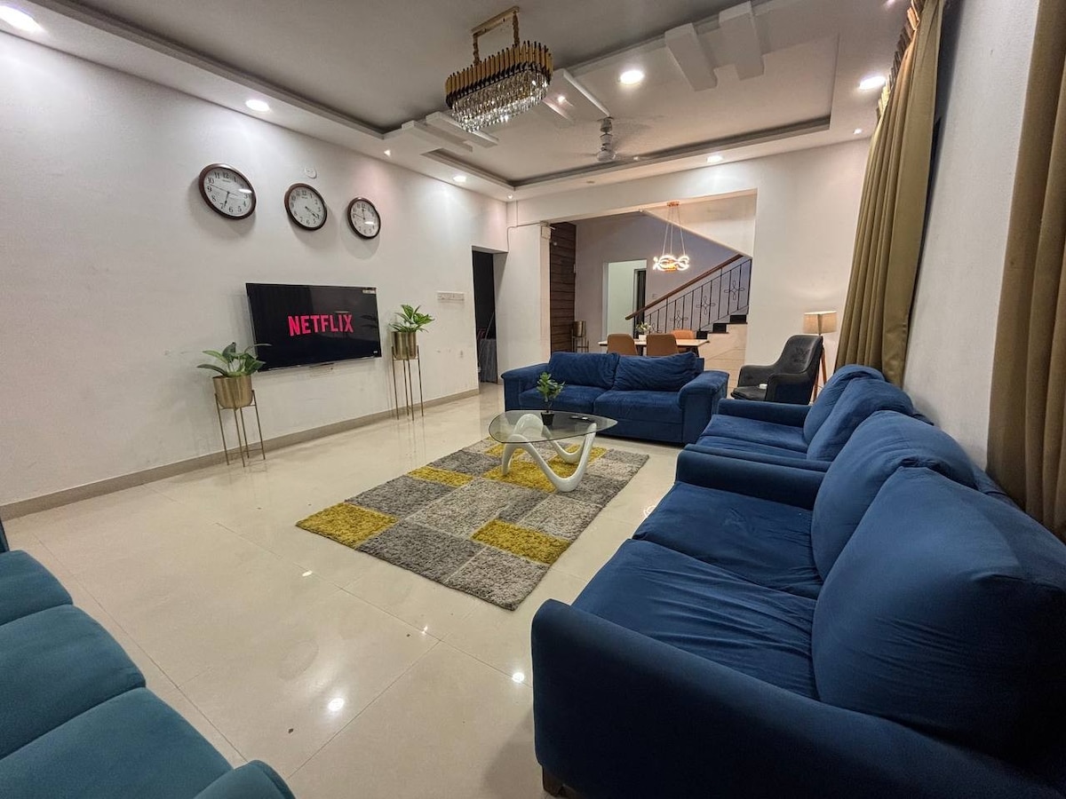 The spacious living room showcases a large blue couch arrangement, comfortably seating multiple guests. A sleek coffee table rests on a patterned rug, while a wall-mounted TV displays Netflix. Elegant lighting and decorative plants enhance the open and inviting atmosphere.