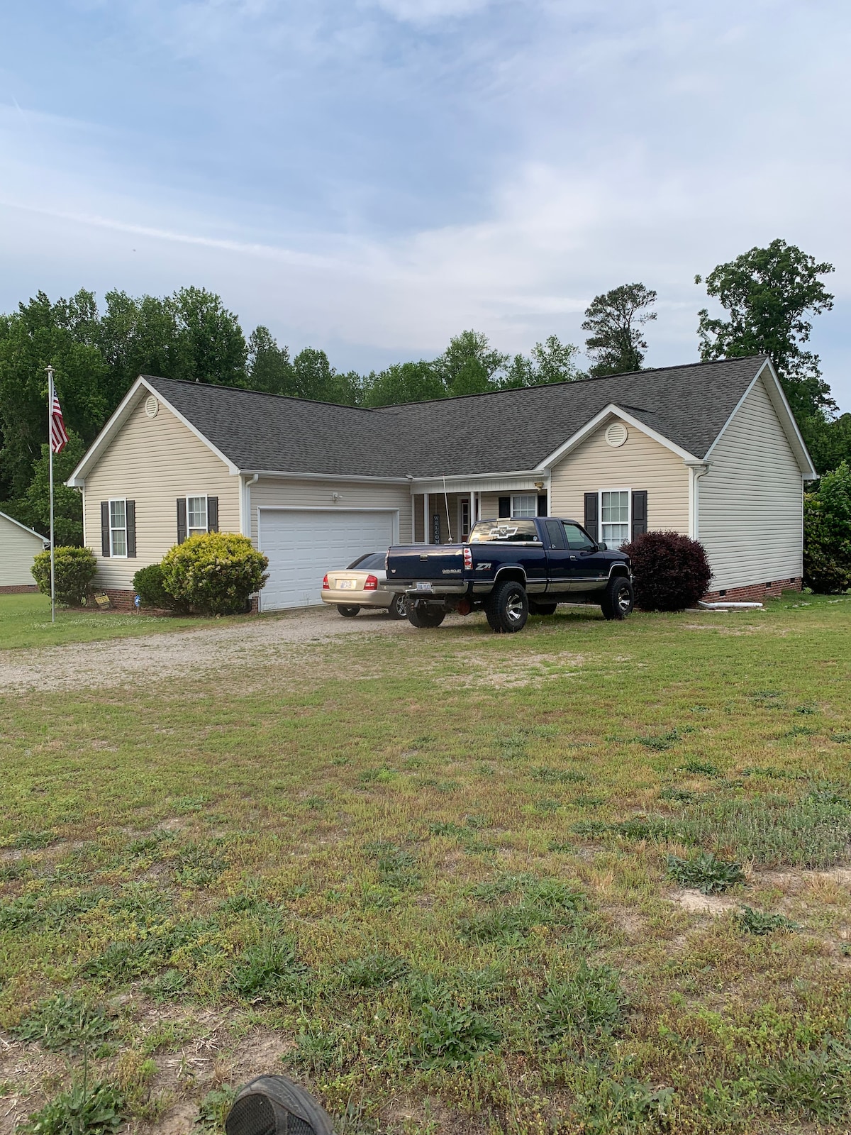 A single-story house is viewed from the front, showcasing a light-colored exterior with a neatly landscaped yard. A black pickup truck is parked in the driveway adjacent to a garage. An American flag is displayed in front of the house, surrounded by greenery.