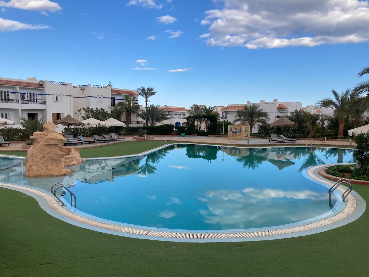 A spacious swimming pool is surrounded by well-maintained green lawns and palm trees. Loungers are arranged around the pool area, while a rock feature adds a natural touch. The sky reflects on the water's surface, creating a serene scene.
