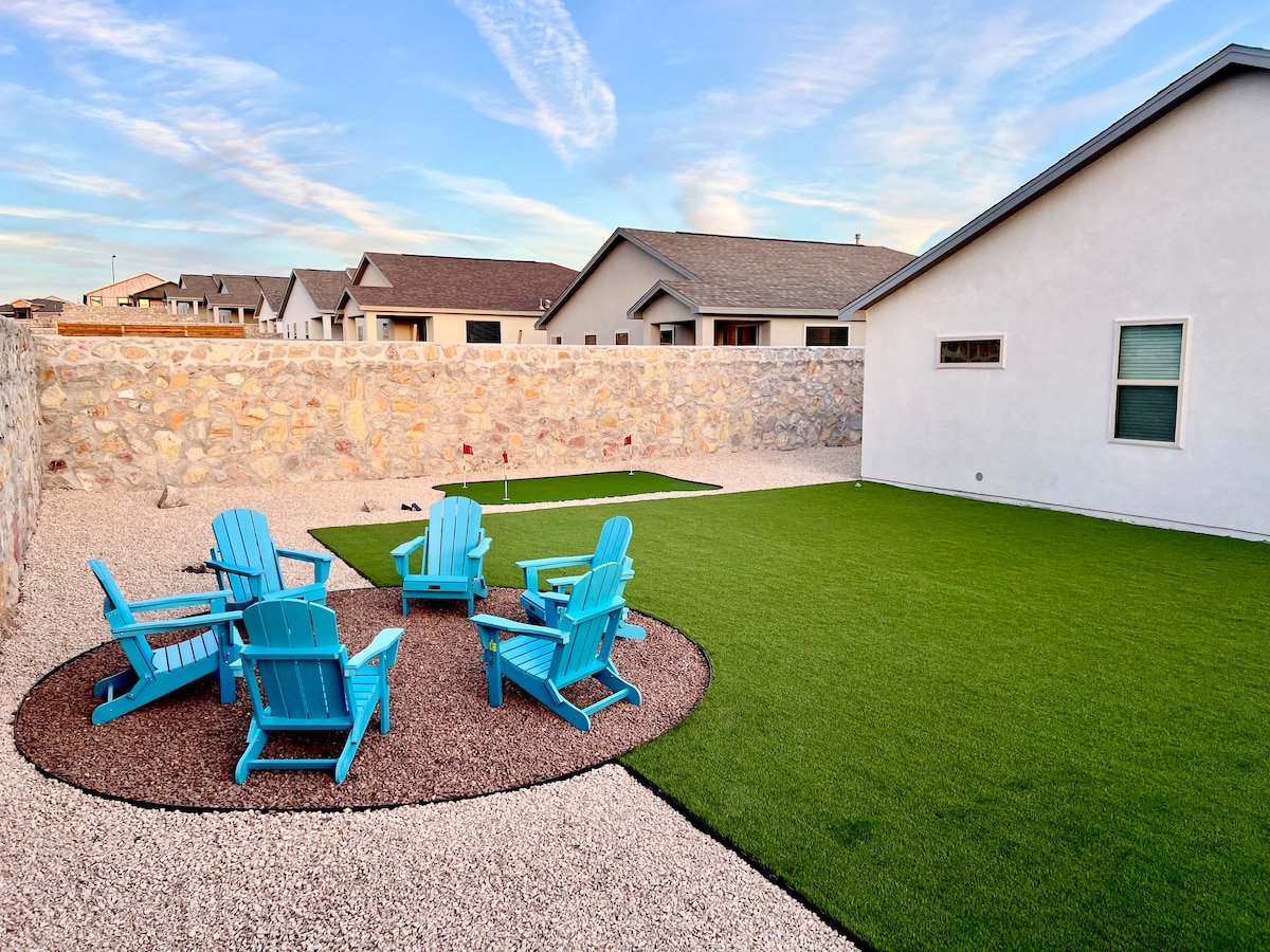 An outdoor space is featured with a well-maintained green lawn surrounded by decorative gravel. Several bright blue Adirondack chairs are arranged in a circular pattern, offering a cozy seating area. Low stone walls provide privacy, with the sky visible in the background.