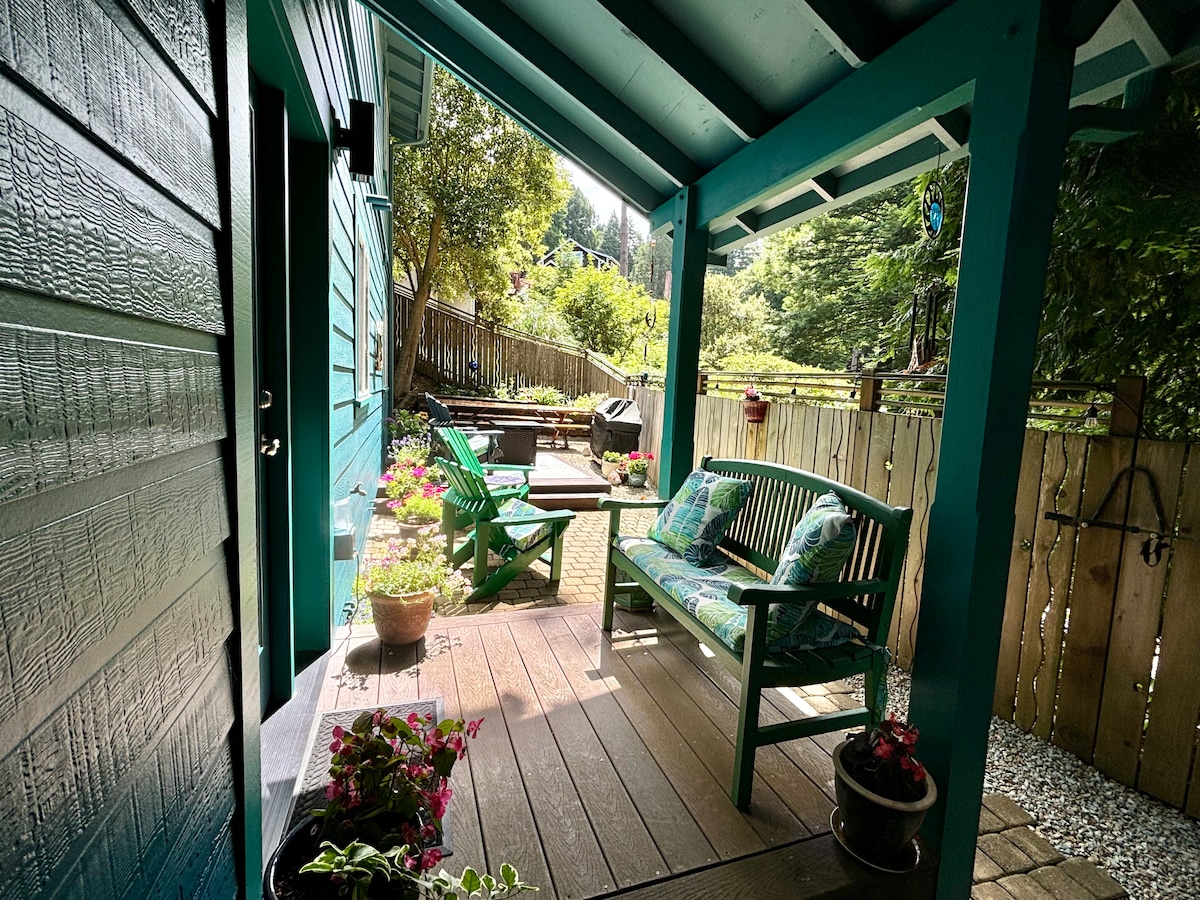 A cozy outdoor patio area features a green bench with cushions, inviting relaxation. Potted plants add color, while a doorway leads to the interior. A privacy fence surrounds the area, complemented by natural greenery in the background.