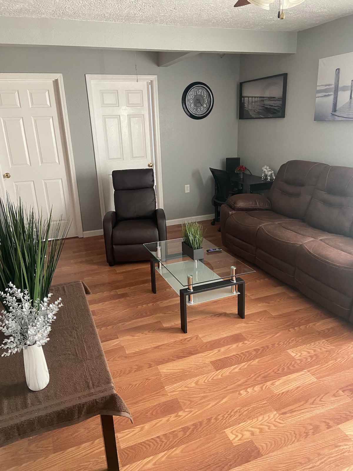 A comfortable living area showcases a brown reclining chair next to a glass coffee table. A dark sofa accommodates seating, and a small table displays a decorative plant. The warm wooden flooring enhances the inviting atmosphere, complemented by neutral wall tones.
