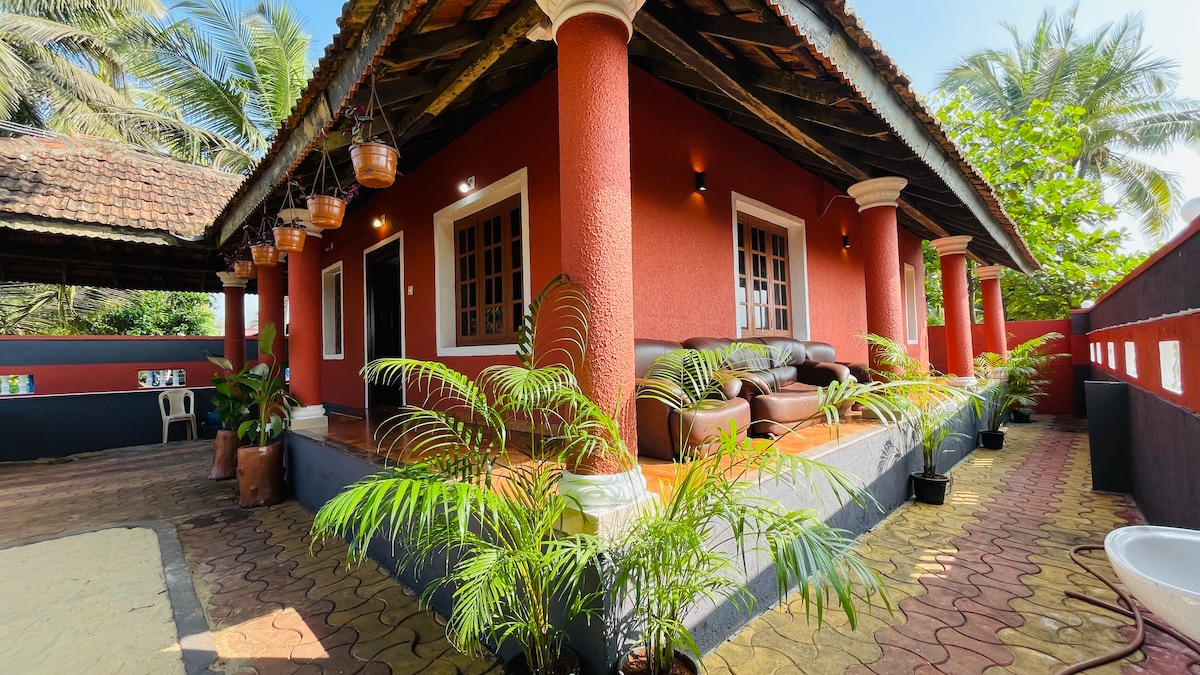 The exterior of the villa features a vibrant red facade with decorative columns. A shaded veranda is visible, adorned with potted plants and a cozy seating area. The lush greenery and palm trees provide a tropical atmosphere, enhancing the inviting appearance of the beachside retreat.