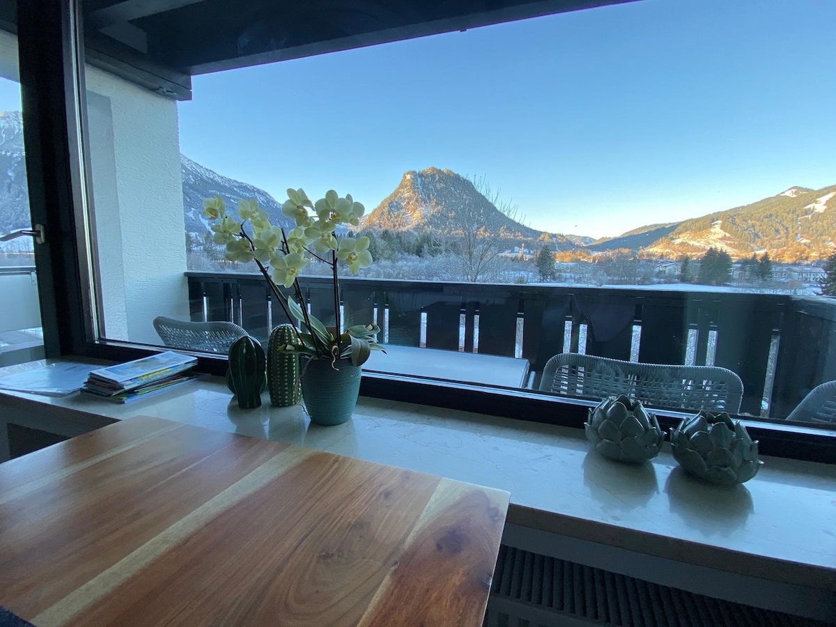 A bright and airy space is created by large windows, offering a mountain view. A wooden table is positioned near the window with potted plants including orchids and succulents adding a touch of greenery. Outdoor seating can be seen on the balcony outside.