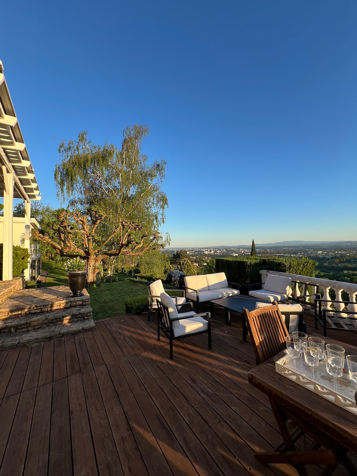 A spacious wooden terrace is furnished with comfortable seating arranged for relaxation, overlooking a vast landscape. The area features a table set for dining, with glasses visible. Lush greenery and a large tree complement the picturesque view of the valley and distant mountains.
