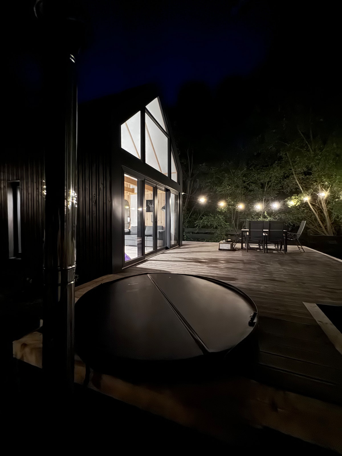 An evening view of the cabin showcases a spacious terrace with outdoor dining furniture and string lights illuminating the space. A hot tub is positioned nearby, nestled against the wooden deck. Large glass doors reflect the warm light from inside the cabin.