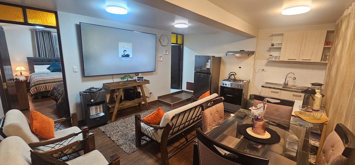 A modern living area features a large flat-screen television mounted on the wall, alongside a dining table with multiple chairs. Comfortable seating with decorative cushions is arranged to create a welcoming space. A well-equipped kitchen is visible in the background, enhancing convenience.