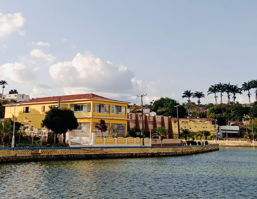 A vibrant yellow house is situated by the water, surrounded by lush greenery and palm trees. The building features a charming red-tiled roof and is complemented by a landscaped area, reflecting a homely atmosphere in a scenic setting.