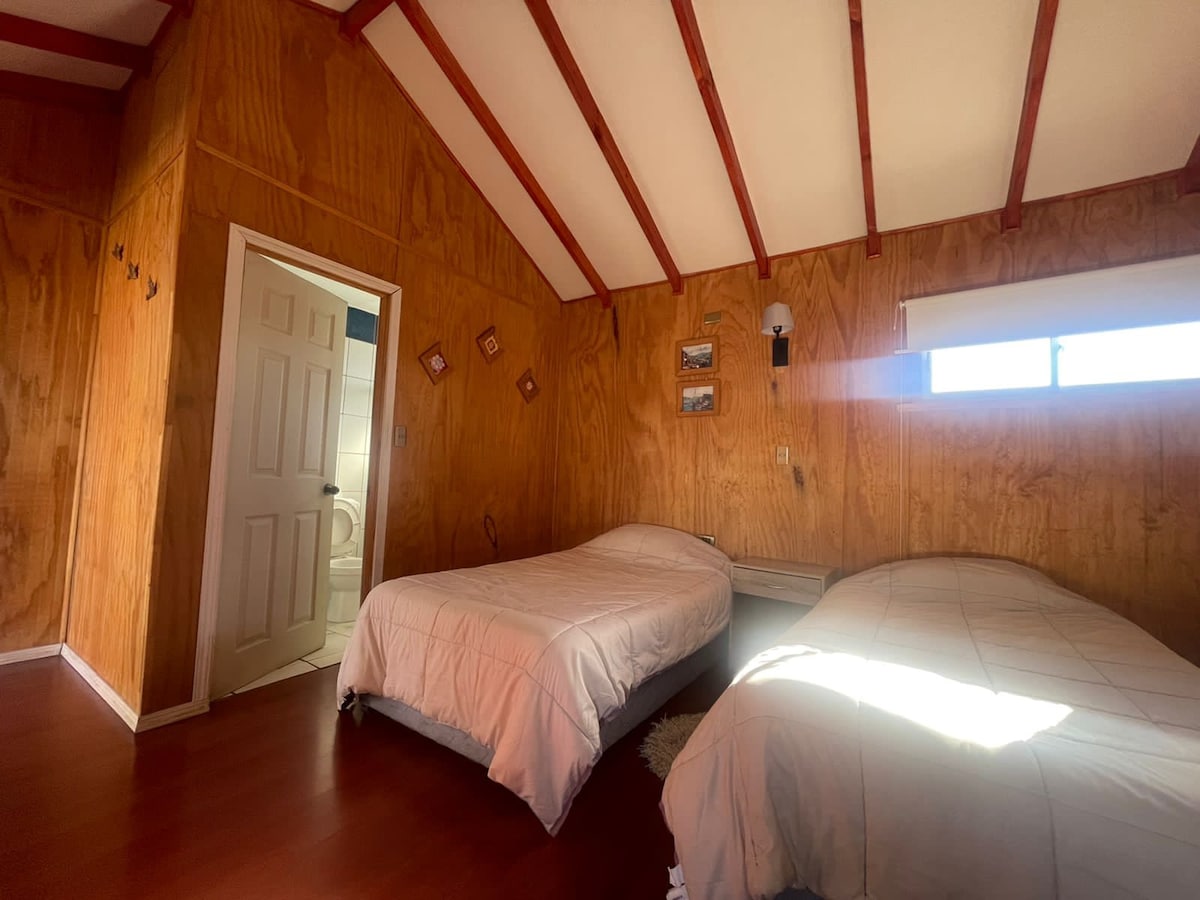 A well-lit sleeping area is furnished with two beds, each covered with light-colored bedding. The walls are adorned with warm wooden panels, and a door leads to a bathroom area. Natural light enters through a horizontal window above the beds.