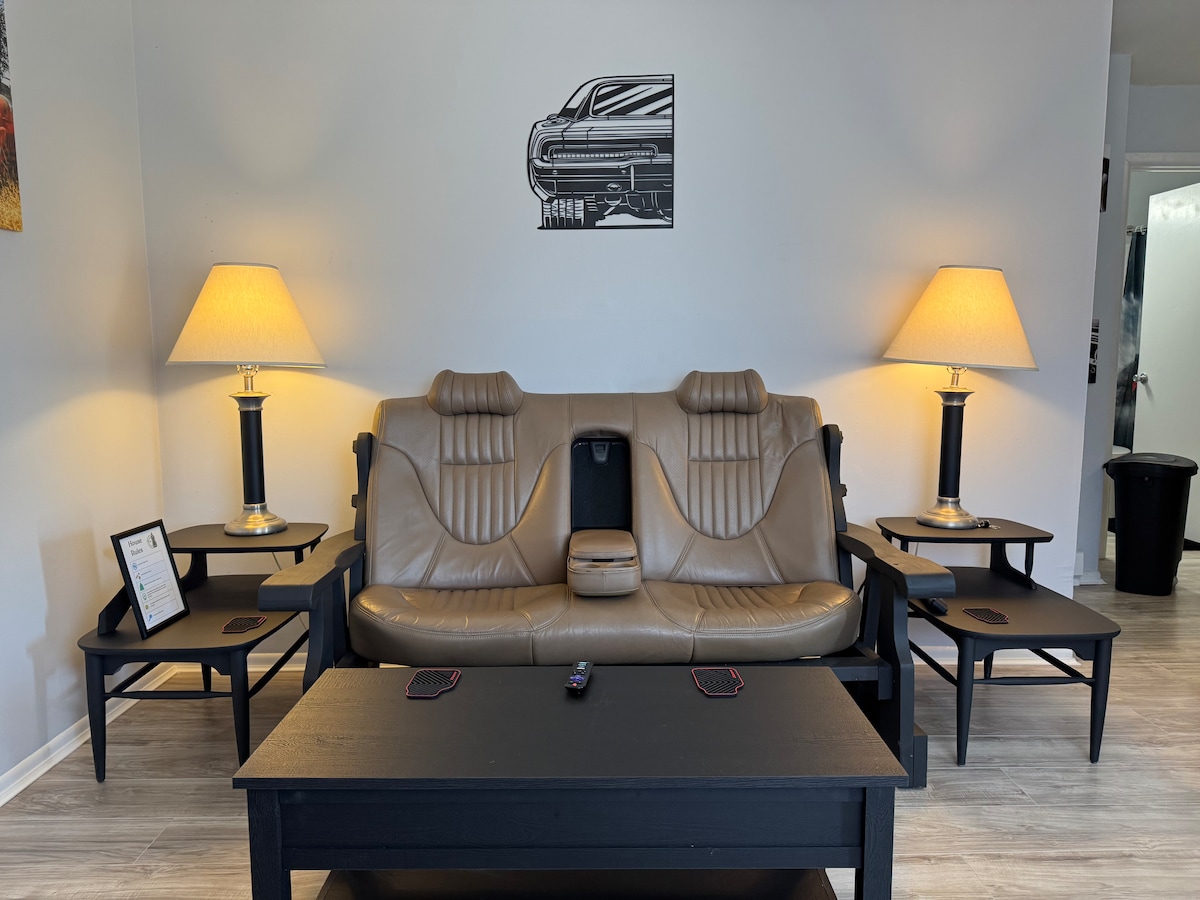 A unique couch crafted from a car seat is positioned in a bright living area. Flanking the couch are two black side tables, each topped with matching lamps that provide soft lighting. A black coffee table sits in front, complemented by a wall art featuring a classic car.