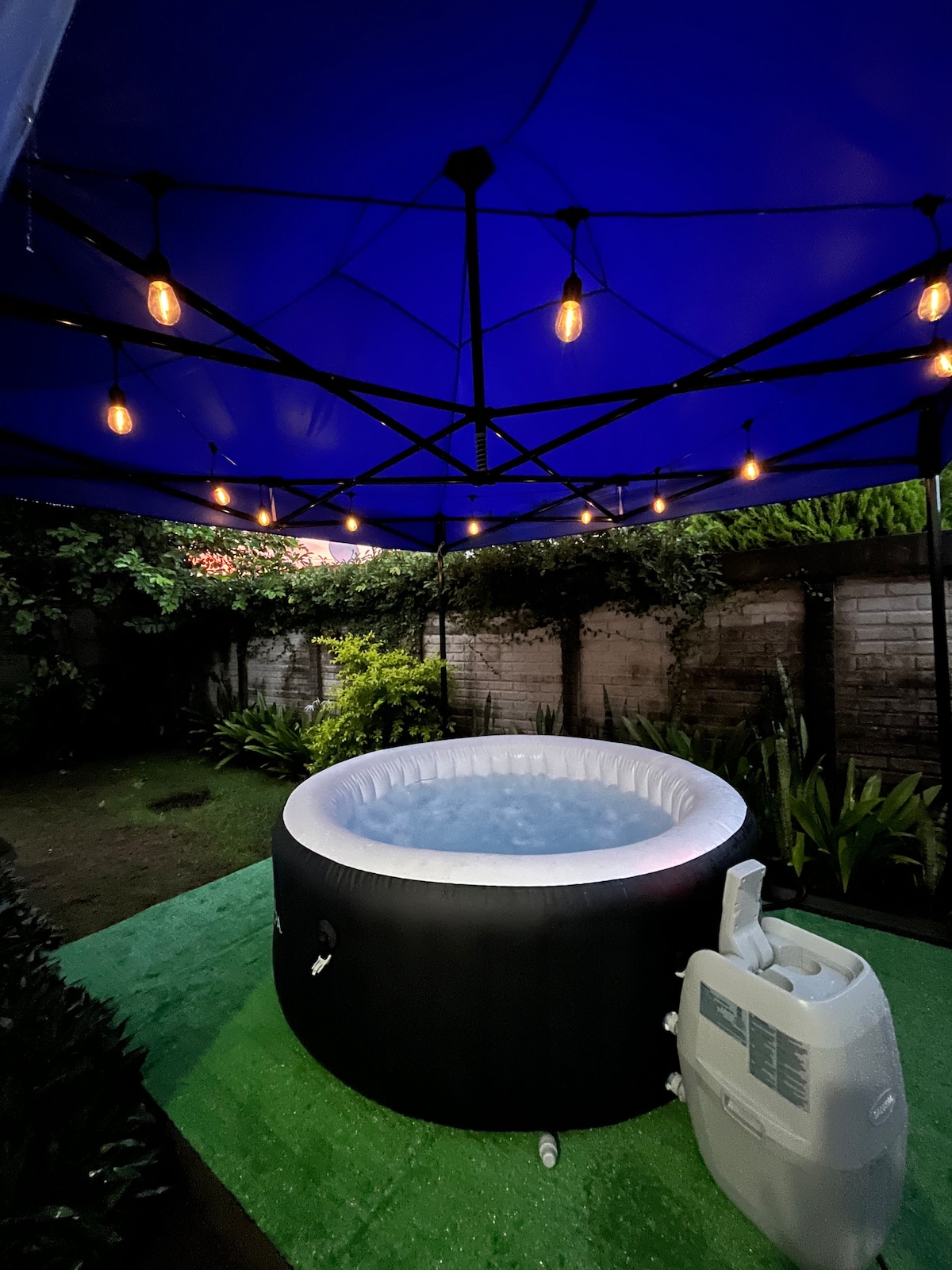An outdoor hot tub is positioned under a blue canopy, surrounded by lush greenery and soft lighting. The tub features a soothing water surface, and a pump is discreetly placed nearby, enhancing the relaxing atmosphere.