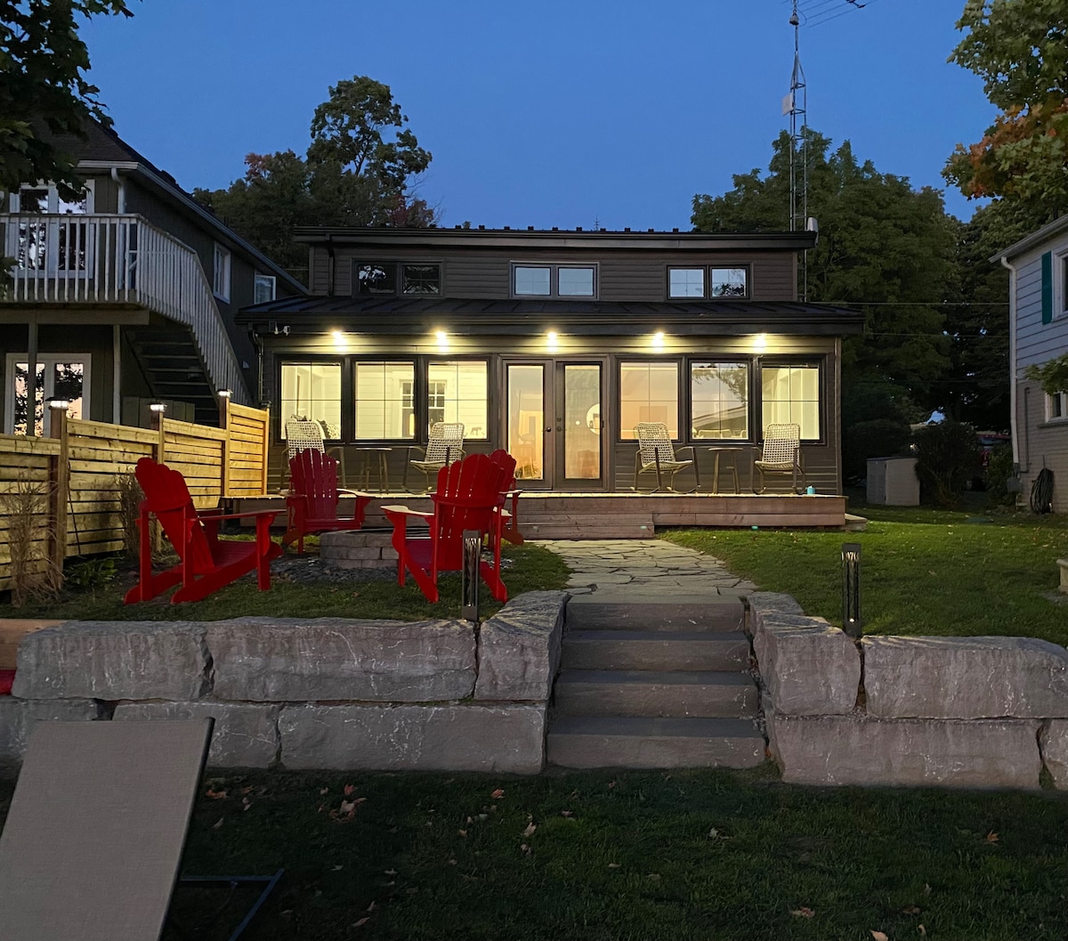The exterior of the cottage is illuminated at dusk, showcasing large glass doors that open to a welcoming outdoor space. Red Adirondack chairs are arranged around a stone-firepit area, leading up to a manicured lawn and a stone pathway.